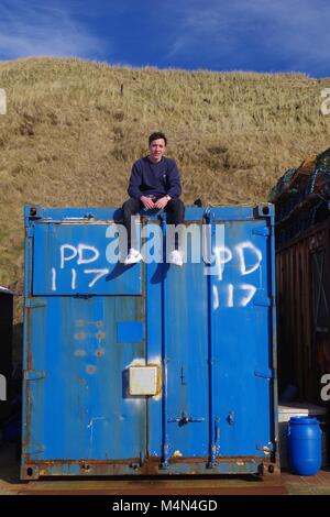 Junge Mann auf einen alten blauen Versandverpackung an Cruden Bay Harbour, Aberdeenshire, Schottland, Großbritannien sitzen. Winter, 2018. Stockfoto