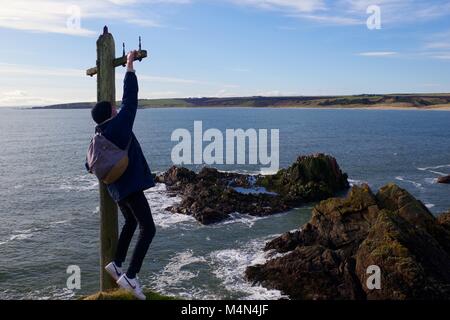 Junge Abenteuerlustige Mann baumelt aus einem alten Telegraphenmast am Rande eines Liff. Cruden Bay, Aberdeenshire, Schottland, Großbritannien. Feb., 2018. Stockfoto