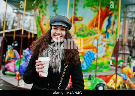 Curly mexikanische Mädchen in Leder und Plastik Tasse Kaffee in der Hand gegen Karussell. Stockfoto