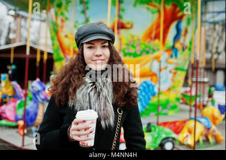 Curly mexikanische Mädchen in Leder und Plastik Tasse Kaffee in der Hand gegen Karussell. Stockfoto