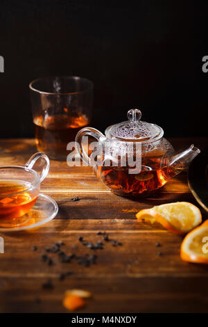 Kleine Teekanne aus Glas mit heißem schwarzem Tee, Becher, Gläser, gepresste Orange Slice auf Holz- Hintergrund. Abendlicht. Vertikale Komposition. Stockfoto