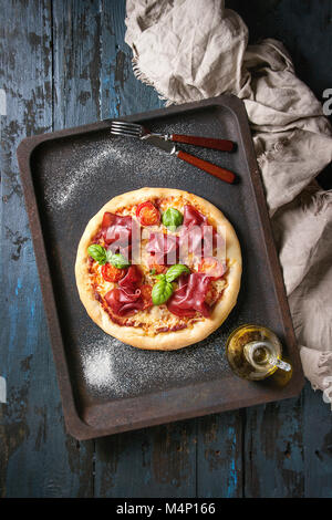 Traditionelle Pizza mit Bresaola, Käse, Tomaten und Basilikum serviert auf alten Backblech mit textilen über Dunkelblau Holz- Hintergrund. Ansicht von oben mit Platz Stockfoto