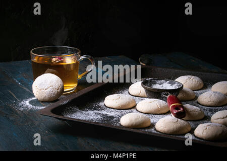 Hausgemachte almond Cookies mit Zucker Pulver, mit Vintage Sieb, die auf alten Backblech und heiße Tasse Tee über Dunkelblau Holztisch. Dunklen rustikalen Stil. Top Stockfoto