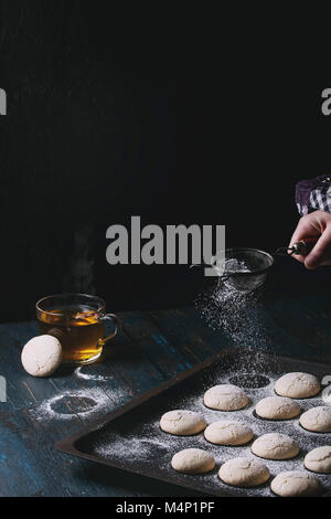 Hausgemachte almond Cookies auf alten Ofen Fach und heiße Tasse Tee über Dunkelblau Holztisch. Prise Zucker Pulver von Vintage Sieb in der Hand des Kindes. Dunkel Stockfoto