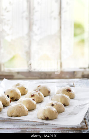 Raw ungebackene Gebäck. Bereit, hausgemachte Ostern traditionelle Hot Cross Buns auf Backpapier über weiße Holztisch mit Fenster im Hintergrund zu backen. Natürliche Stockfoto