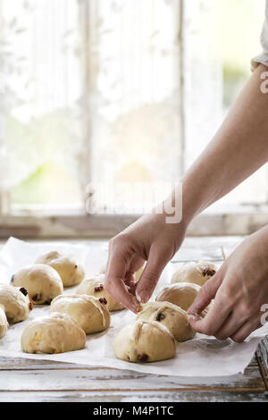 Raw ungebackene Gebäck. Bereit, hausgemachte Ostern traditionelle Hot Cross Buns auf Backpapier über weiße Holztisch zu backen. Im Hintergrund angezeigt. Weibliche Hände Stockfoto