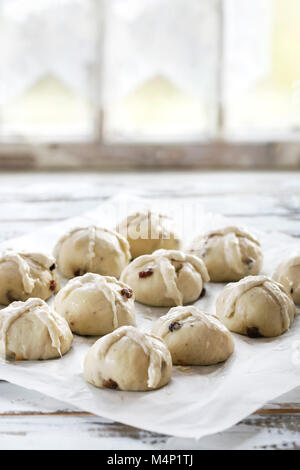 Raw ungebackene Gebäck. Bereit, hausgemachte Ostern traditionelle Hot Cross Buns auf Backpapier über weiße Holztisch mit Fenster im Hintergrund zu backen. Natürliche Stockfoto