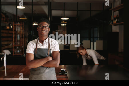 Mann in seinen Kaffee shop mit gekreuzten Armen, während Frau im Hintergrund arbeitet. Jungen Unternehmern ihre Coffee Shop verwalten. Stockfoto