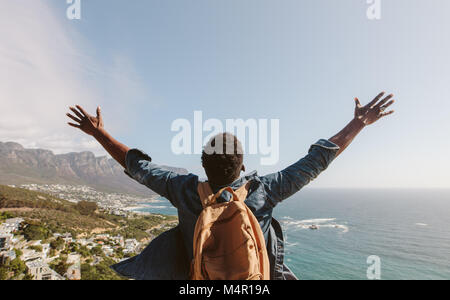 Ansicht der Rückseite des jungen Mann mit Rucksack im Freien stand mit ausgebreiteten Armen gegen Seascape. Man genießt die Aussicht von der Spitze des Berges. Stockfoto