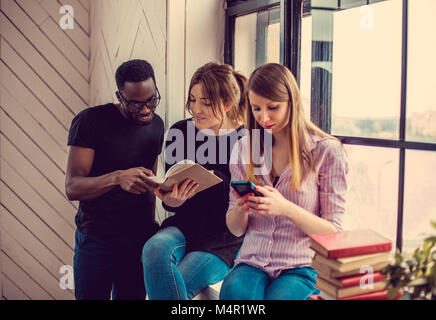 Afrikanischer amerikanischer Mann und zwei kaukasischen Frauen ein Buch lesen in der Nähe der Fenster. Stockfoto