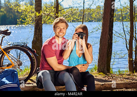 Positive sportliche Paar entspannend in der Nähe des Flusses nach der Fahrt mit dem Fahrrad. Weibliche Aufnehmen von Bildern auf eine kompakte Digitalkamera. Stockfoto