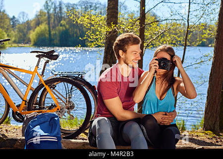 Positive sportliche Paar entspannend in der Nähe des Flusses nach der Fahrt mit dem Fahrrad. Weibliche Aufnehmen von Bildern auf eine kompakte Digitalkamera. Stockfoto