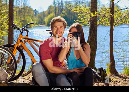 Positive sportliche Paar entspannend in der Nähe des Flusses nach der Fahrt mit dem Fahrrad. Weibliche Aufnehmen von Bildern auf eine kompakte Digitalkamera. Stockfoto