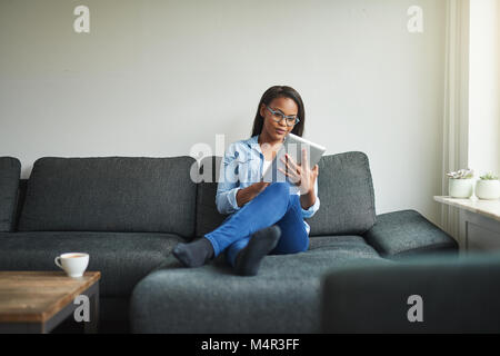 Junge afrikanische Frau Surfen online mit einem digitalen Tablet beim Sitzen auf dem Sofa im Wohnzimmer Stockfoto