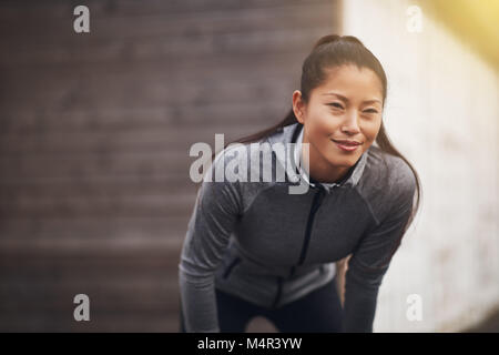 Lächelnde junge asiatische Frau stehend außerhalb übung Kleidung tragen, mit den Händen auf den Knien die Vorbereitung für einen Lauf zu gehen Stockfoto