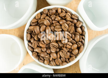 Leicht geröstet braun Kaffeebohnen durch weiße Tassen auf einen hölzernen Tisch umgeben Stockfoto