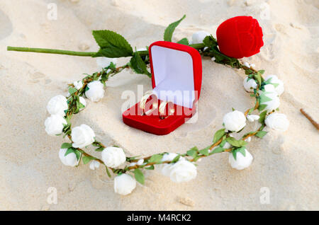 Red Box mit Hochzeit Ringe in der Mitte von einem Kranz aus weißen Blumen auf dem Sand und eine kleine Rose im Hintergrund Stockfoto