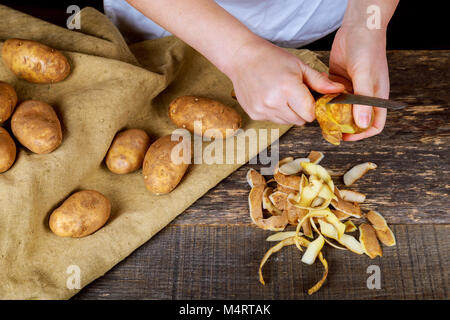 Frau Hände schälen Kartoffeln, Peelings auf Holz Schneidebrett. Drei sauberen Kartoffeln auf dem Teller sauber die Kartoffeln kochen. Stockfoto