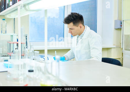 Profil anzeigen von hübscher junger Forscher Gummihandschuhe zu tragen und weißes Fell am Schreibtisch sitzen und sich Notizen während der Durchführung von Experiment in der moder Stockfoto