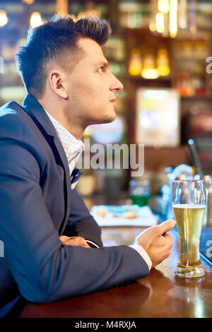 Profil anzeigen von gutaussehenden jungen Mann mit stilvollen Anzug Sitzen an der Theke nach einem harten Arbeitstag, trinken Bier und beobachtete Rugby-spiel Stockfoto
