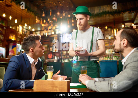 Hübscher junger Mann mit stilvollen Anzug sitzen im Cafe Tabelle mit seinem Kollegen und um Während bärtigen Kellner tragen Kobold Kostüm taki Stockfoto