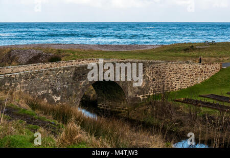 Alte Stein humpback Bridge acorss kleiner Fluss, Nethermill, Aberdeenshire, Schottland, Großbritannien, mit Kiesstrand und Nordsee Stockfoto