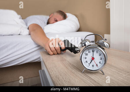 Junger Mann auf dem Bett liegend Halten der Waffe zu schießen Wecker Stockfoto