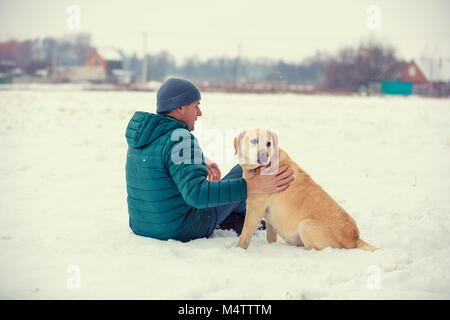 Ein Mann mit einem Labrador Retriever Hund sitzt im Schnee auf ein Feld vor dem Dorf Stockfoto