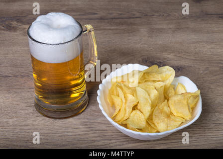 Bier Pint und knusprige Kartoffelchips auf einer hölzernen Tisch. Leicht alkoholischen Bier im Glas mug und Haufen von Fett gebratene Chips in einer weißen Schüssel. Stockfoto