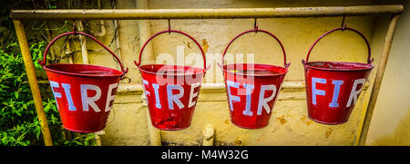 Eine Reihe von vier roten Eimer mit der Bezeichnung "Feuer" in weißen Buchstaben für das Sammeln von Wasser ein Feuer gegen gelbe Wand mit grünen Sträuchern und Reben zu begießen. Stockfoto
