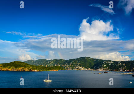 Charlotte Amalie, St. Thomas, USVI. Segelboot und Hügel von Waterfront Hafen mit Regenbogen durch Wolken im blauen Himmel emporragen. Stockfoto