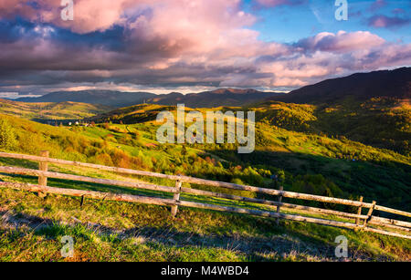 Wunderschönen Abend im bergigen Land. Zaun entlang der grasigen Hang. bergrücken unter der violetten Wolken an einem blauen Himmel. schöne Landschaft im fi Stockfoto