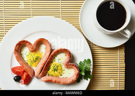 Rührei in der Form von Herzen auf einer weißen Platte mit Wurst, Tomaten, grüne und Kaffee mit Zitrone. Stockfoto