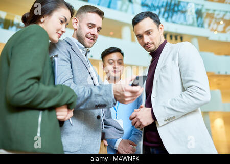 Low Angle Porträt des modernen Geschäftsmann holding Smartphone etwas anzeigen zu Gruppe der Kolleginnen und Kollegen in Halle der Bürogebäude stehend Stockfoto