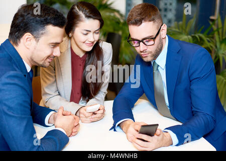 Portrait von drei jungen Geschäftsleuten mit Smartphones am Tisch sitzen im Café, auf der Suche nach Bildschirm und lächelnd Stockfoto