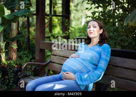 Junge schwangere Frau sitzt im Park auf der Bank. Stockfoto