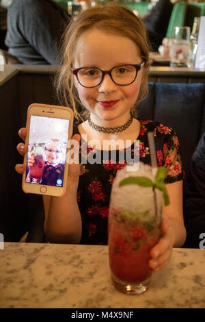 8-Jähriges Mädchen zeigen snapchat auf Ihrem iPhone Smartphone während des Sitzens in einer Bar trinken einen alkoholfreien Cocktail Stockfoto