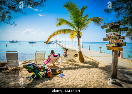 Internationale Ziele Wegweiser am Pier nächsten Lounges und Palmen am Strand mit Blauer Leguan statue Chaise in Rum Point Club, Grand Cayman. Stockfoto