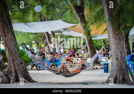 Junger Mann ruht auf einer Hängematte aufhängen von großen Bäumen neben Picknick Tischen Rum Point Club Attraktion im Grand Caymans. Stockfoto