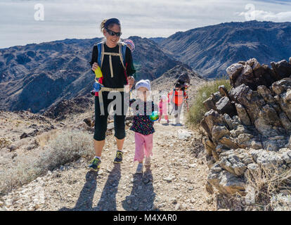 Fit und Jugendlich Mutter Wanderungen mit junge Tochter und Baby in einem Rucksack Carrier auf der Fortynine trail Palms Oase in Joshua Tree National Park. Stockfoto