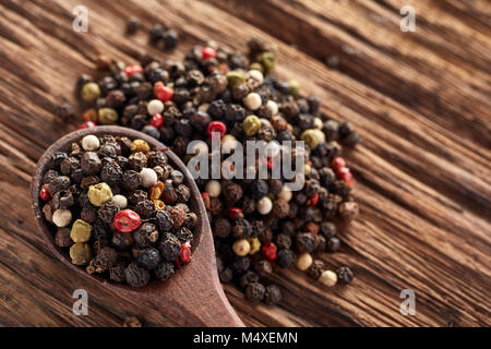 Ansicht von oben in einem hölzernen Löffel voll von schwarzen, roten, gelben, weißen Pfefferkörner auf dunklem Hintergrund. Es isa Haufen von piment Samen hinter sich. Stockfoto