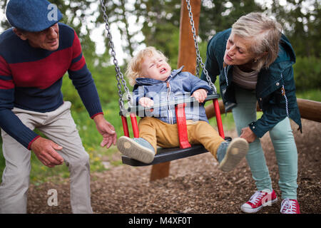 Senior Paar mit Enkel auf dem Spielplatz. Kleiner Junge auf der Schaukel, weinen. Stockfoto