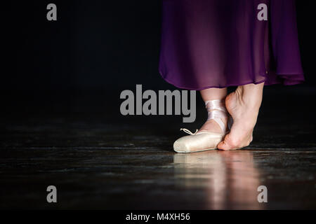 Elegante Ballerina posiert im Studio auf dem dunklen Hintergrund. Sie steht auf der linken Zehe und hält das rechte Bein auf dem Boden. Mädchen trägt einen beigefarbenen Po Stockfoto