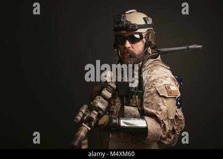 Brutaler Mann in der militärischen Wüste einheitliche und Body Armour steht in einem Kampf Rack und hält sein Gewehr auf schwarzem Hintergrund. Die bärtige Spieler in Th Stockfoto