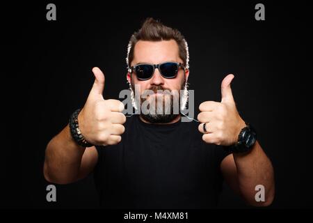 Ein schöner, starker Mann mit einem stilvollen Frisur und Bart zeigt zwei Daumen hoch im Studio auf einem schwarzen Hintergrund. Kopieren mit Platz für Ihren Text. Stockfoto