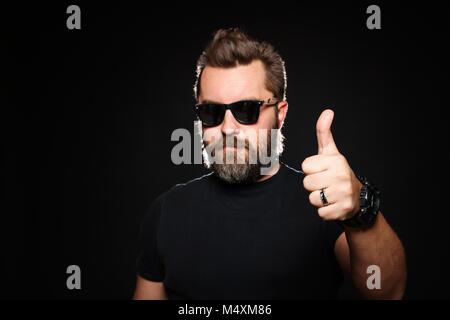 Ein schöner, starker Mann mit einem stilvollen Frisur und Bart zeigt Daumen hoch, im Studio auf einem schwarzen Hintergrund. Kopieren mit Platz für Ihren Text. Stockfoto