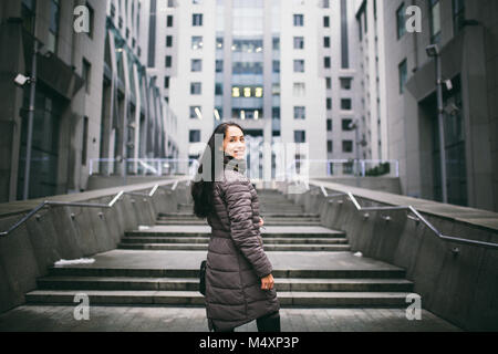 Porträt einer Frau, die auf einem Thema. Junge kaukasier brunette Mädchen in langen Jacke, Mantel schwarzes Leder Tasche steht auf Business Center backgroun Stockfoto