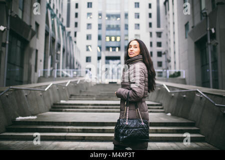 Porträt einer Frau, die auf einem Thema. Junge kaukasier brunette Mädchen in langen Jacke, Mantel schwarzes Leder Tasche steht auf Business Center backgroun Stockfoto