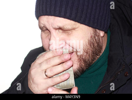 Ein bärtiger Mann in einer blauen Daunenjacke und einem strickmütze Husten, über seinen Mund mit einem Taschentuch Stockfoto