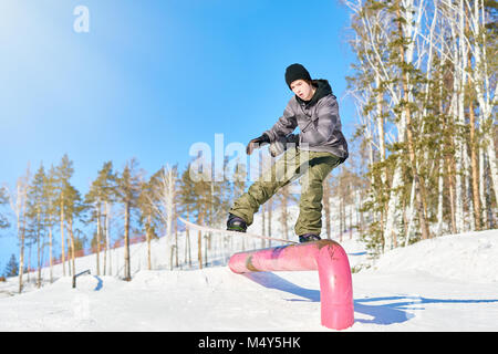 In voller Länge Porträt des jungen Mannes, Durchführung von Snowboard stunt Hineinrutschen Metallgeländer im Sonnenlicht am Ski Resort, kopieren Raum Stockfoto